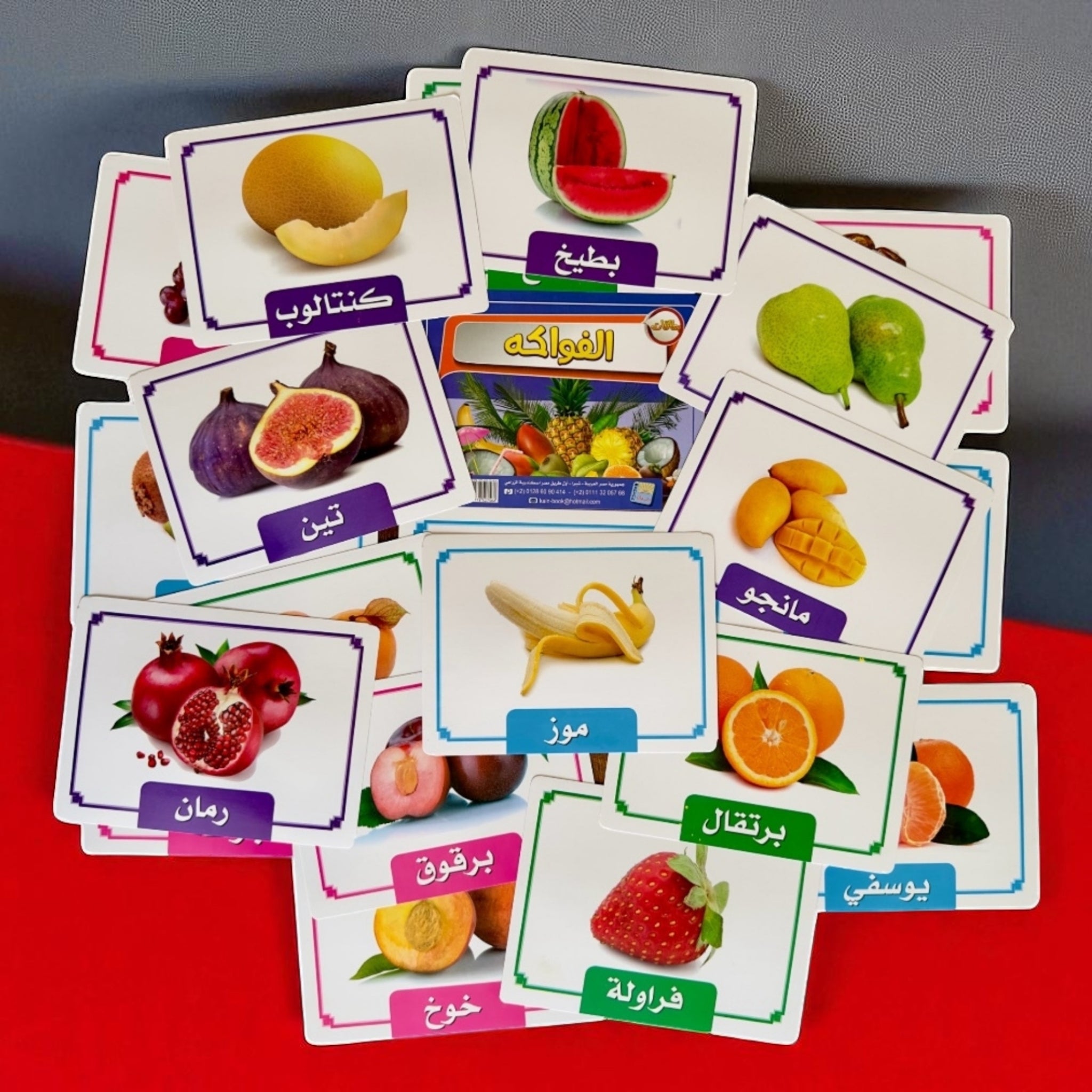 Educational cards with images of fruits and their names in Arabic on a red surface.
